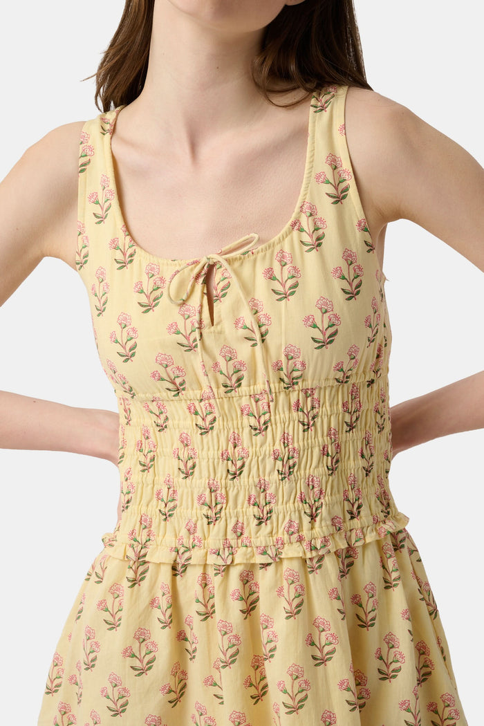 A young woman models the Roller Rabbit Rosa Ditsy Floral Penny Dress, a light yellow sundress with a floral pattern, gathered bodice, and gently flared skirt, set against a plain white background.