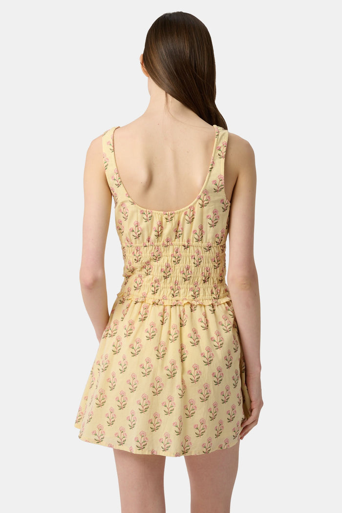 A young woman models the Roller Rabbit Rosa Ditsy Floral Penny Dress, a light yellow sundress with a floral pattern, gathered bodice, and gently flared skirt, set against a plain white background.