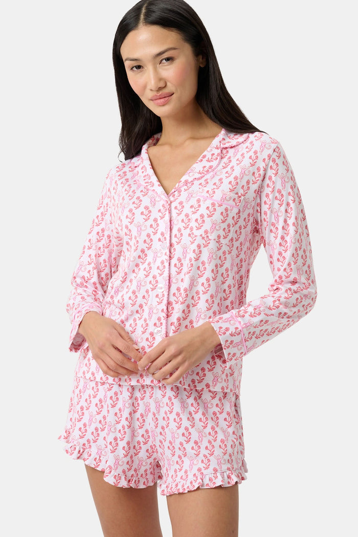 Wearing the Roller Rabbit Rosa Ditsy Monkey Hayden Pajamas—a pink and white set with a small anchor print, long-sleeve button-up top, and ruffled shorts—a person stands against a plain light background, smiling slightly.