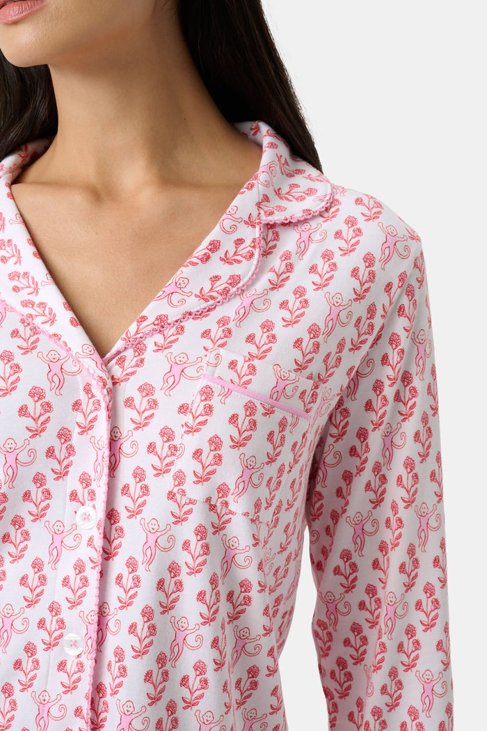 Wearing the Roller Rabbit Rosa Ditsy Monkey Hayden Pajamas—a pink and white set with a small anchor print, long-sleeve button-up top, and ruffled shorts—a person stands against a plain light background, smiling slightly.