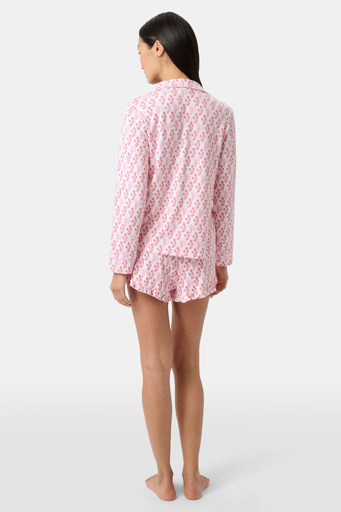 Wearing the Roller Rabbit Rosa Ditsy Monkey Hayden Pajamas—a pink and white set with a small anchor print, long-sleeve button-up top, and ruffled shorts—a person stands against a plain light background, smiling slightly.