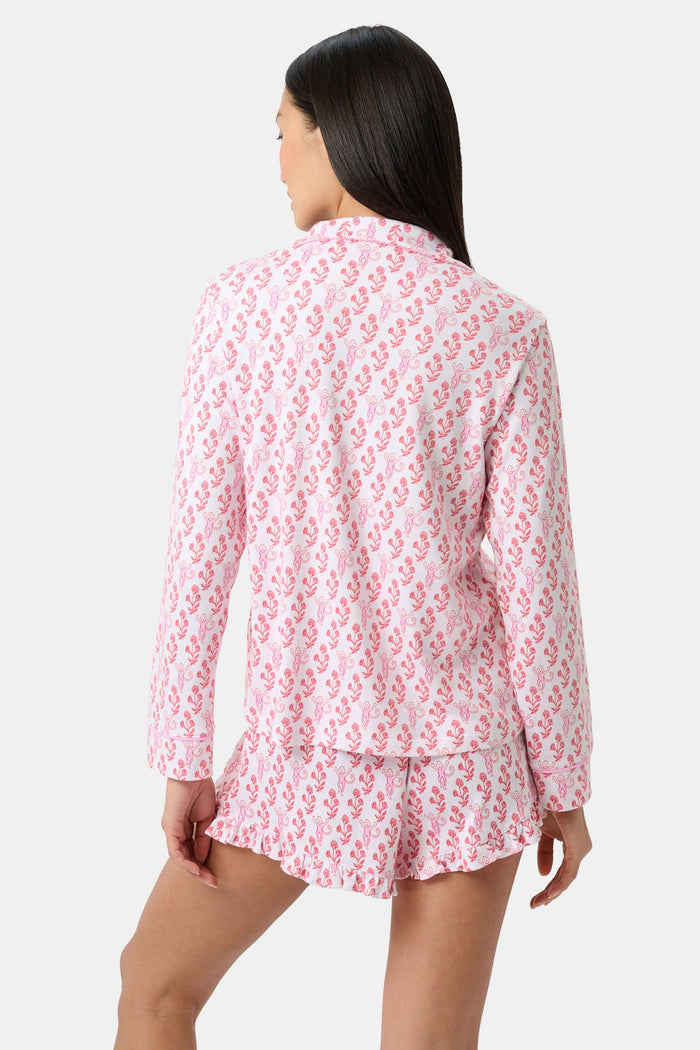 Wearing the Roller Rabbit Rosa Ditsy Monkey Hayden Pajamas—a pink and white set with a small anchor print, long-sleeve button-up top, and ruffled shorts—a person stands against a plain light background, smiling slightly.