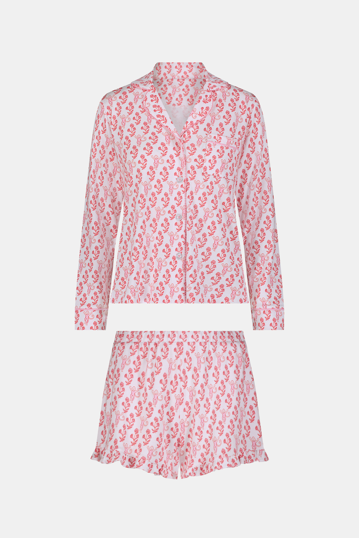Wearing the Roller Rabbit Rosa Ditsy Monkey Hayden Pajamas—a pink and white set with a small anchor print, long-sleeve button-up top, and ruffled shorts—a person stands against a plain light background, smiling slightly.