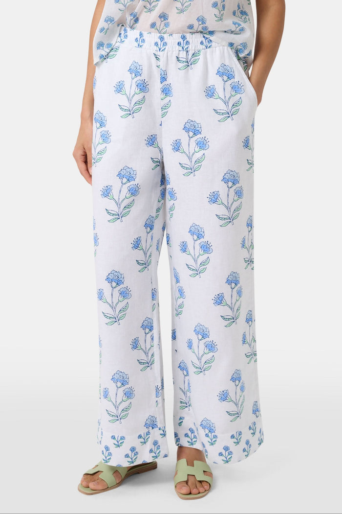 A person wears the Roller Rabbit Rosa Floral Kaylee Pant—high-waisted white linen pants with blue floral print and pockets—paired with an off-white top and green sandals, standing against a plain light background.