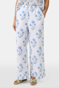 A person wears the Roller Rabbit Rosa Floral Kaylee Pant—high-waisted white linen pants with blue floral print and pockets—paired with an off-white top and green sandals, standing against a plain light background.