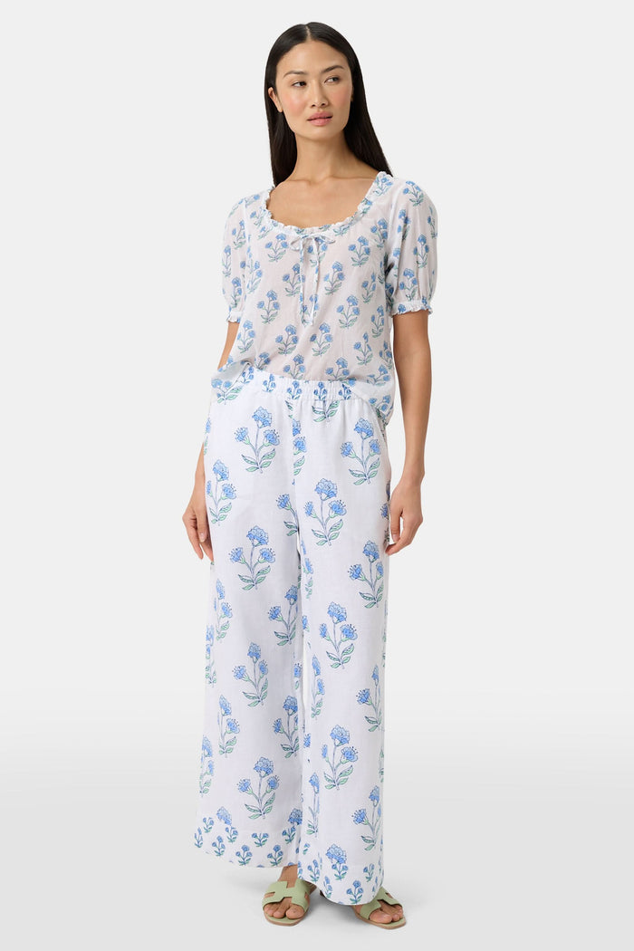 A person wears the Roller Rabbit Rosa Floral Kaylee Pant—high-waisted white linen pants with blue floral print and pockets—paired with an off-white top and green sandals, standing against a plain light background.