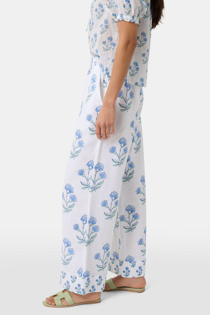A person wears the Roller Rabbit Rosa Floral Kaylee Pant—high-waisted white linen pants with blue floral print and pockets—paired with an off-white top and green sandals, standing against a plain light background.