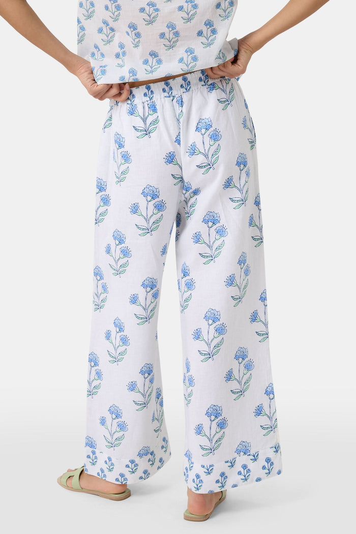 A person wears the Roller Rabbit Rosa Floral Kaylee Pant—high-waisted white linen pants with blue floral print and pockets—paired with an off-white top and green sandals, standing against a plain light background.