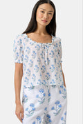 A woman with long dark hair wears the Roller Rabbit Rosa Floral Quincy Top, a cotton voile blouse with puffed sleeves, ruffled tie neckline, and matching blue flower patterns as part of a coordinating set.