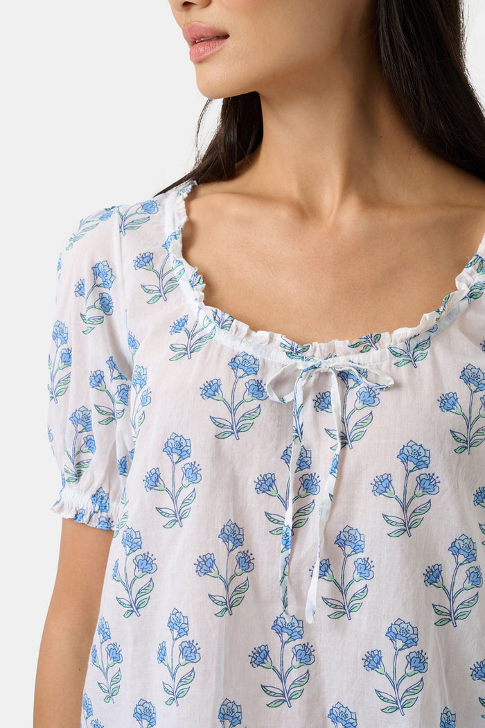 A woman with long dark hair wears the Roller Rabbit Rosa Floral Quincy Top, a cotton voile blouse with puffed sleeves, ruffled tie neckline, and matching blue flower patterns as part of a coordinating set.