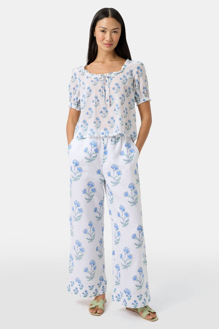 A woman with long dark hair wears the Roller Rabbit Rosa Floral Quincy Top, a cotton voile blouse with puffed sleeves, ruffled tie neckline, and matching blue flower patterns as part of a coordinating set.