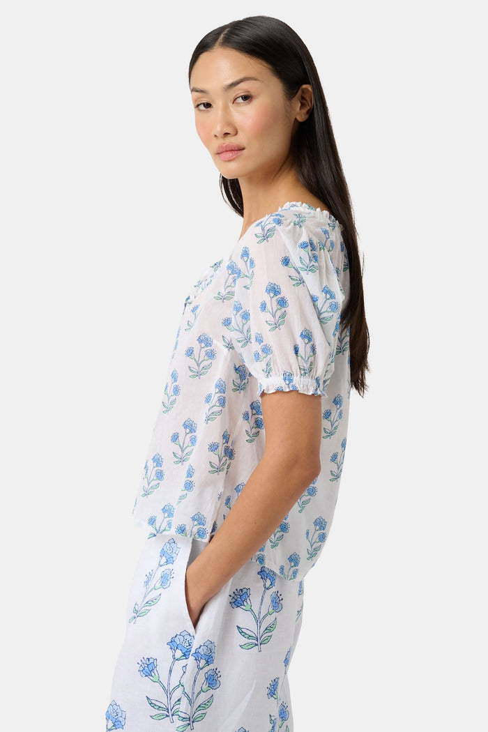 A woman with long dark hair wears the Roller Rabbit Rosa Floral Quincy Top, a cotton voile blouse with puffed sleeves, ruffled tie neckline, and matching blue flower patterns as part of a coordinating set.
