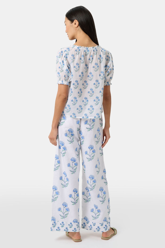 A woman with long dark hair wears the Roller Rabbit Rosa Floral Quincy Top, a cotton voile blouse with puffed sleeves, ruffled tie neckline, and matching blue flower patterns as part of a coordinating set.