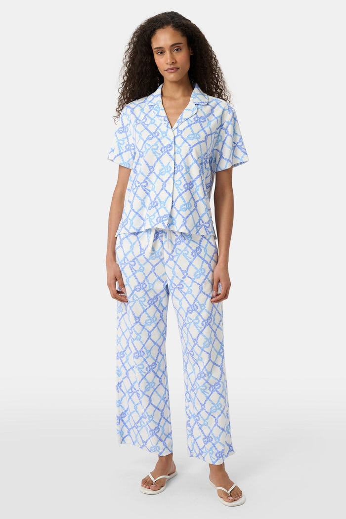 A person wears the Roller Rabbit Seaside Knots Mara Pant, featuring a tie waist and wide legs in loose white fabric with a light blue geometric chain pattern, paired with a matching top and white sandals against a plain white background.