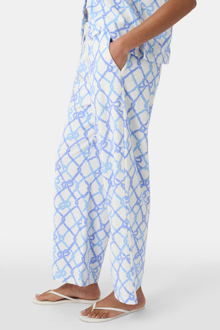 A person wears the Roller Rabbit Seaside Knots Mara Pant, featuring a tie waist and wide legs in loose white fabric with a light blue geometric chain pattern, paired with a matching top and white sandals against a plain white background.