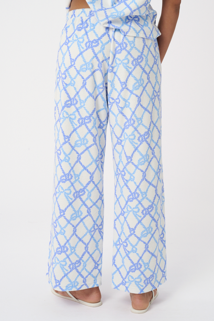 A person wears the Roller Rabbit Seaside Knots Mara Pant, featuring a tie waist and wide legs in loose white fabric with a light blue geometric chain pattern, paired with a matching top and white sandals against a plain white background.