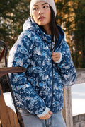 A woman in the Roller Rabbit Ski Toile Reversible Puffer Jacket with a white scenic print, a beige knit beanie, and jeans stands outdoors by a wooden fence amidst snow and trees.
