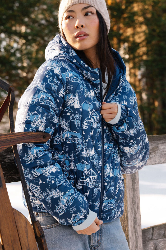 A woman in the Roller Rabbit Ski Toile Reversible Puffer Jacket with a white scenic print, a beige knit beanie, and jeans stands outdoors by a wooden fence amidst snow and trees.