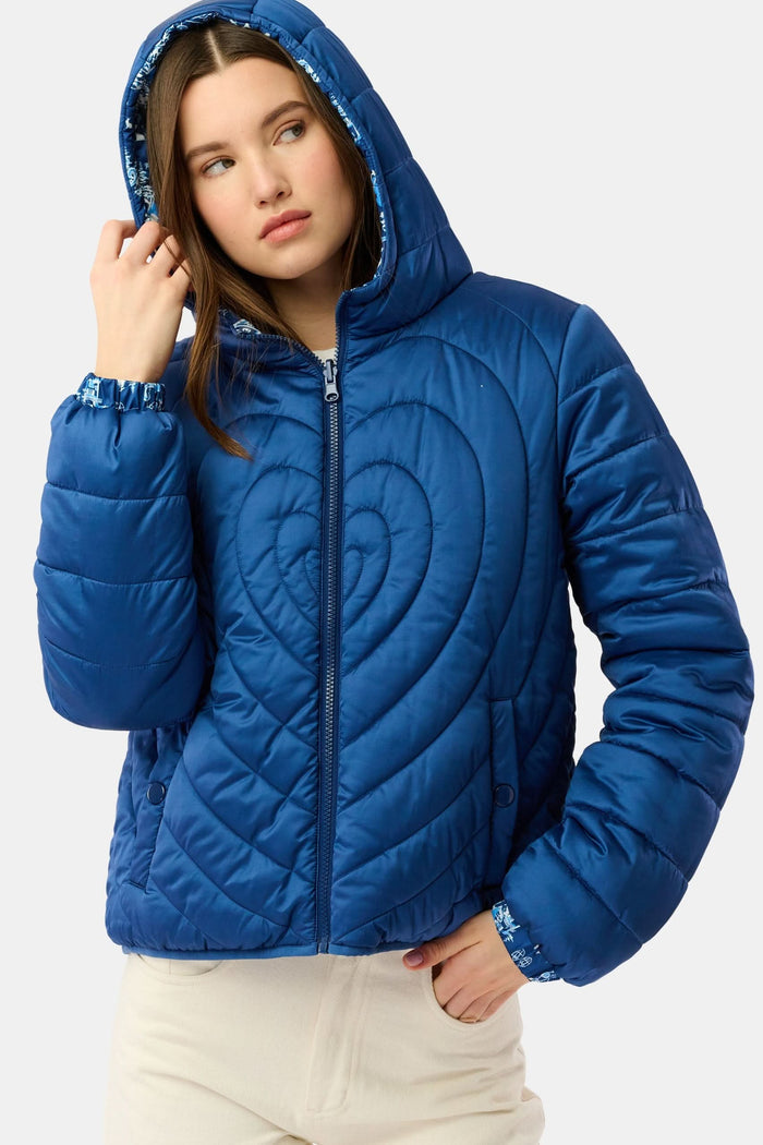 A woman in the Roller Rabbit Ski Toile Reversible Puffer Jacket with a white scenic print, a beige knit beanie, and jeans stands outdoors by a wooden fence amidst snow and trees.