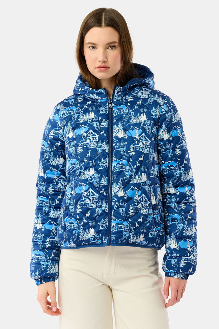 A woman in the Roller Rabbit Ski Toile Reversible Puffer Jacket with a white scenic print, a beige knit beanie, and jeans stands outdoors by a wooden fence amidst snow and trees.