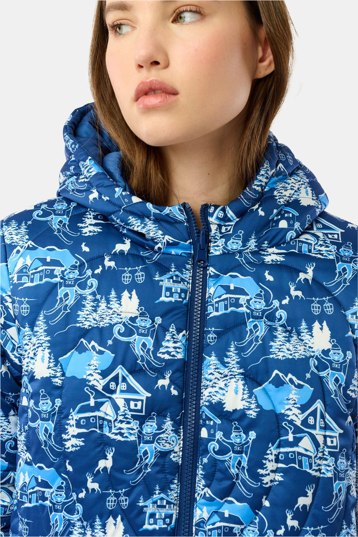 A woman in the Roller Rabbit Ski Toile Reversible Puffer Jacket with a white scenic print, a beige knit beanie, and jeans stands outdoors by a wooden fence amidst snow and trees.