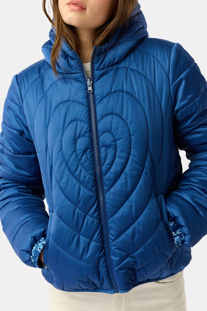 A woman in the Roller Rabbit Ski Toile Reversible Puffer Jacket with a white scenic print, a beige knit beanie, and jeans stands outdoors by a wooden fence amidst snow and trees.