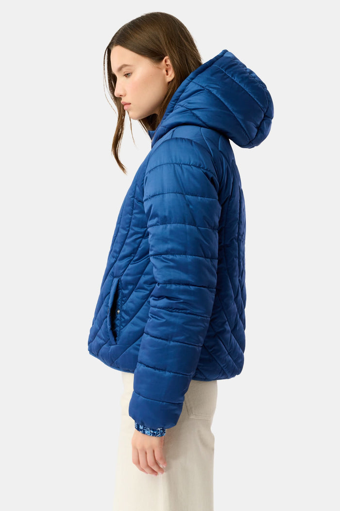 A woman in the Roller Rabbit Ski Toile Reversible Puffer Jacket with a white scenic print, a beige knit beanie, and jeans stands outdoors by a wooden fence amidst snow and trees.
