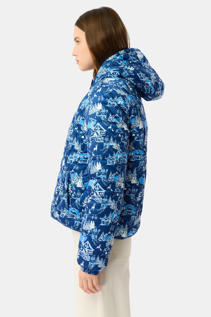 A woman in the Roller Rabbit Ski Toile Reversible Puffer Jacket with a white scenic print, a beige knit beanie, and jeans stands outdoors by a wooden fence amidst snow and trees.