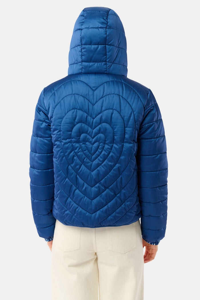 A woman in the Roller Rabbit Ski Toile Reversible Puffer Jacket with a white scenic print, a beige knit beanie, and jeans stands outdoors by a wooden fence amidst snow and trees.