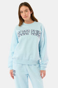 A woman stands against a plain background wearing the Roller Rabbit Sleep Club Andie Sweatshirt, a light blue top with bold retro varsity-style SLEEP CLUB lettering across the chest, paired with matching sweatpants.