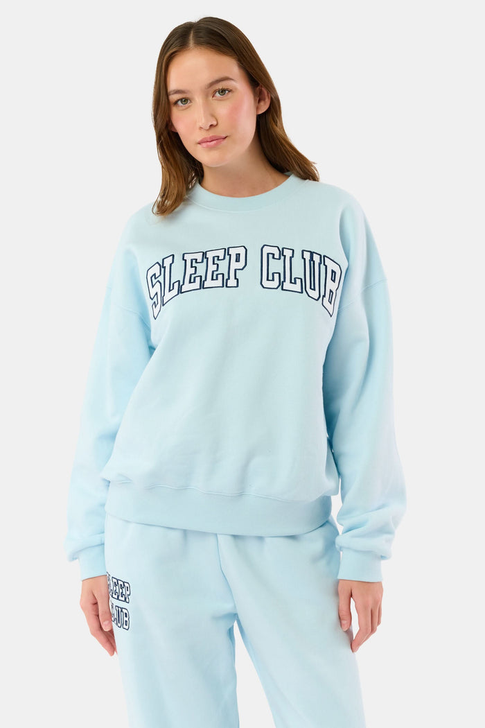 A woman stands against a plain background wearing the Roller Rabbit Sleep Club Andie Sweatshirt, a light blue top with bold retro varsity-style SLEEP CLUB lettering across the chest, paired with matching sweatpants.