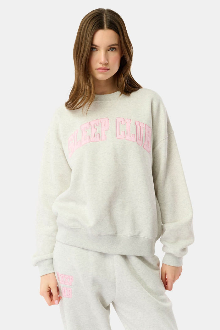 A person relaxes on a bed with pink bedding, wearing the Roller Rabbit Sleep Club Andie Sweatshirt and holding a white mug, smiling softly at the camera.