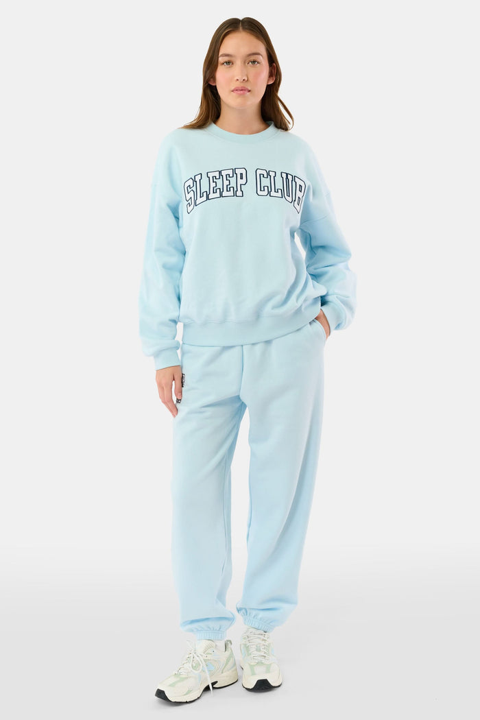 A woman stands against a plain background wearing the Roller Rabbit Sleep Club Andie Sweatshirt, a light blue top with bold retro varsity-style SLEEP CLUB lettering across the chest, paired with matching sweatpants.