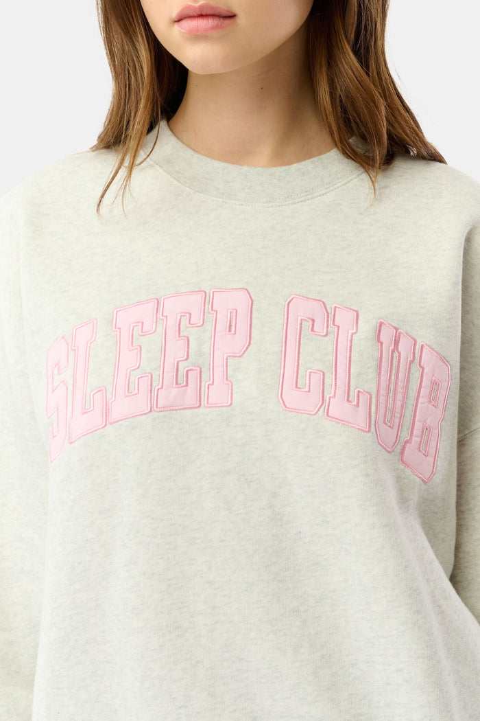A person relaxes on a bed with pink bedding, wearing the Roller Rabbit Sleep Club Andie Sweatshirt and holding a white mug, smiling softly at the camera.