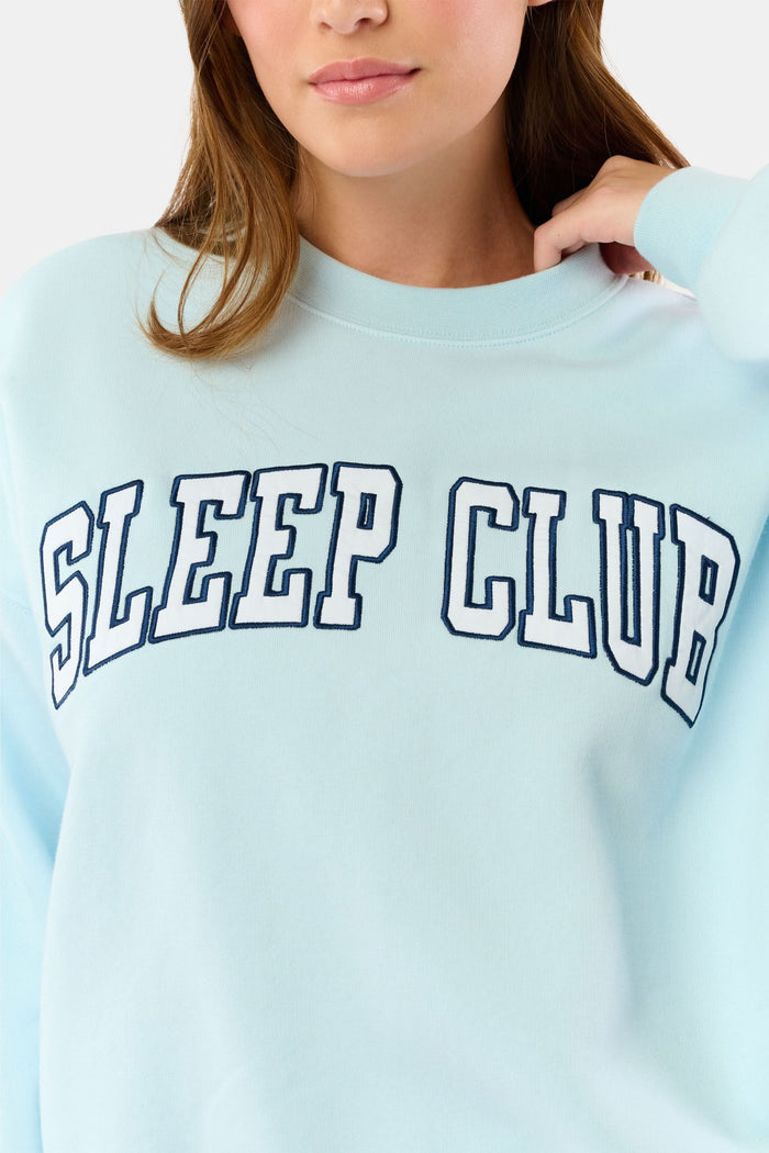 A woman stands against a plain background wearing the Roller Rabbit Sleep Club Andie Sweatshirt, a light blue top with bold retro varsity-style SLEEP CLUB lettering across the chest, paired with matching sweatpants.