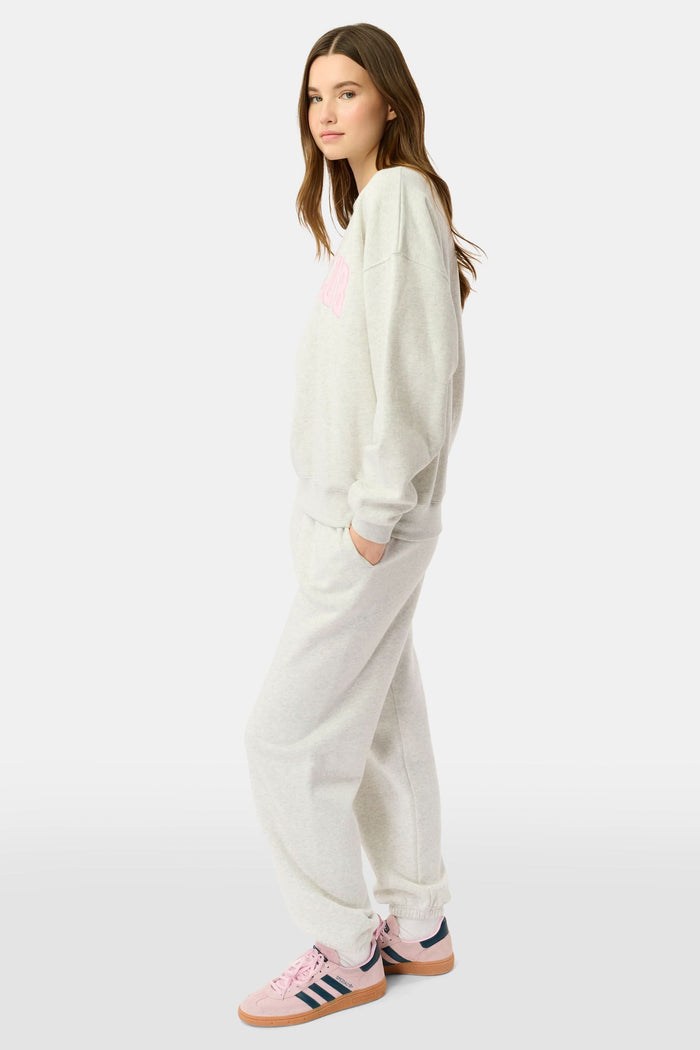 A person relaxes on a bed with pink bedding, wearing the Roller Rabbit Sleep Club Andie Sweatshirt and holding a white mug, smiling softly at the camera.