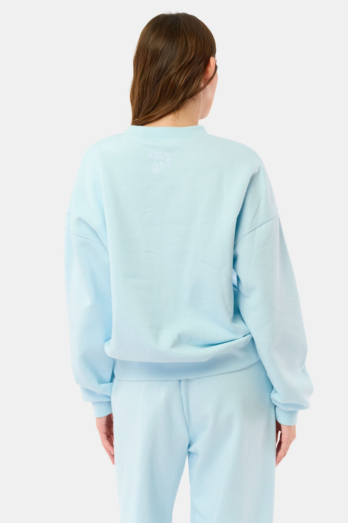 A woman stands against a plain background wearing the Roller Rabbit Sleep Club Andie Sweatshirt, a light blue top with bold retro varsity-style SLEEP CLUB lettering across the chest, paired with matching sweatpants.