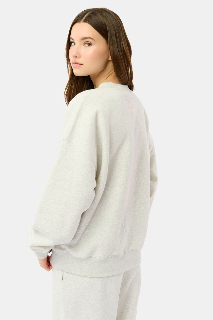 A person relaxes on a bed with pink bedding, wearing the Roller Rabbit Sleep Club Andie Sweatshirt and holding a white mug, smiling softly at the camera.