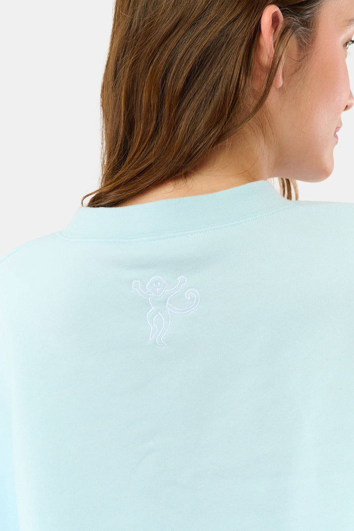 A woman stands against a plain background wearing the Roller Rabbit Sleep Club Andie Sweatshirt, a light blue top with bold retro varsity-style SLEEP CLUB lettering across the chest, paired with matching sweatpants.
