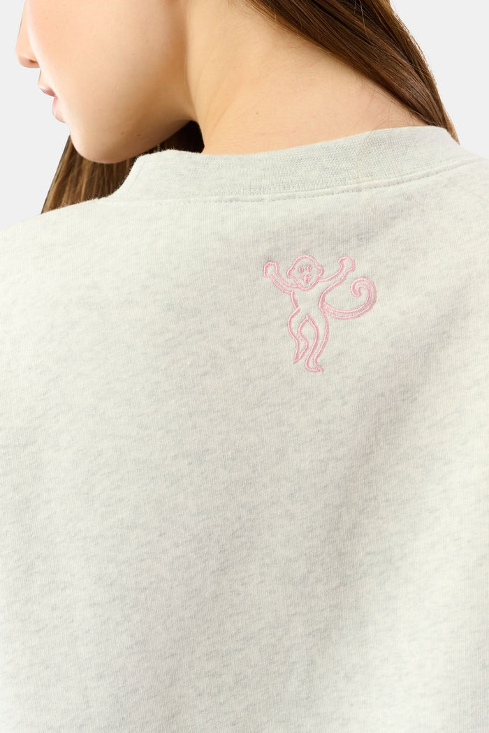 A person relaxes on a bed with pink bedding, wearing the Roller Rabbit Sleep Club Andie Sweatshirt and holding a white mug, smiling softly at the camera.