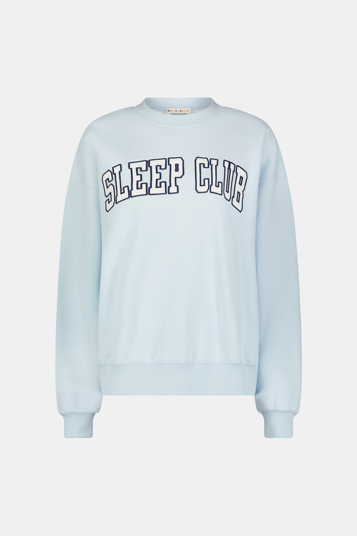 A woman stands against a plain background wearing the Roller Rabbit Sleep Club Andie Sweatshirt, a light blue top with bold retro varsity-style SLEEP CLUB lettering across the chest, paired with matching sweatpants.