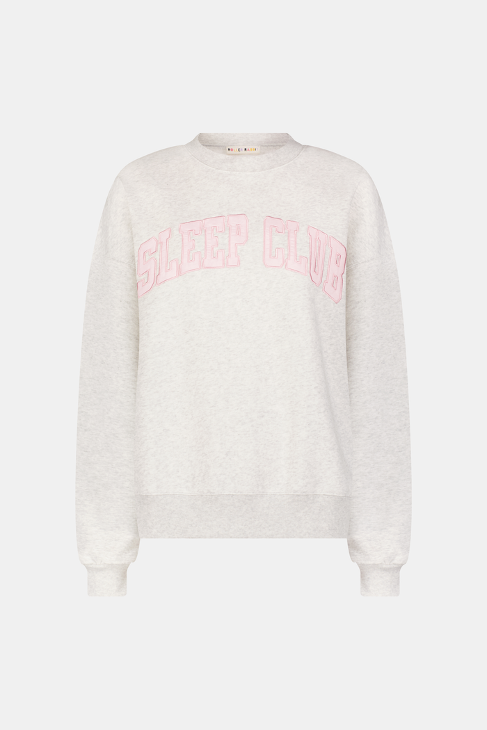 A person relaxes on a bed with pink bedding, wearing the Roller Rabbit Sleep Club Andie Sweatshirt and holding a white mug, smiling softly at the camera.