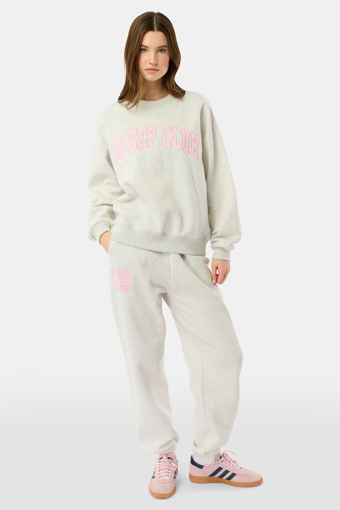 A woman stands against a white background wearing light gray Roller Rabbit Sleep Club Bea Sweatpants and a matching sweatshirt with "SLEEP CLUB" in pink, styled with pink and black Adidas sneakers.