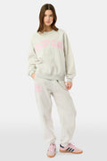A woman stands against a white background wearing light gray Roller Rabbit Sleep Club Bea Sweatpants and a matching sweatshirt with "SLEEP CLUB" in pink, styled with pink and black Adidas sneakers.