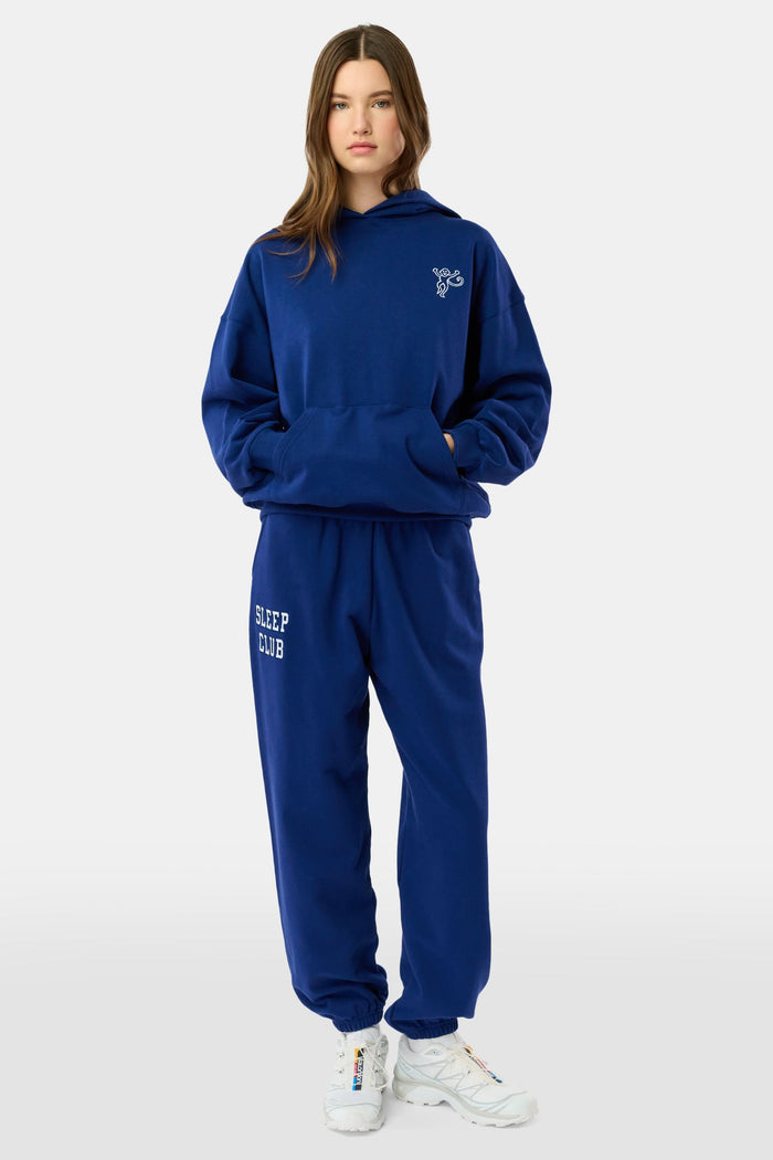 A young woman faces forward in a matching blue hoodie and Roller Rabbit Sleep Club Bea Sweatpants with "SLEEP CLUB" printed in white, paired with white sneakers. The hoodie has a small white graphic.