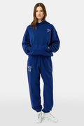 A young woman faces forward in a matching blue hoodie and Roller Rabbit Sleep Club Bea Sweatpants with "SLEEP CLUB" printed in white, paired with white sneakers. The hoodie has a small white graphic.