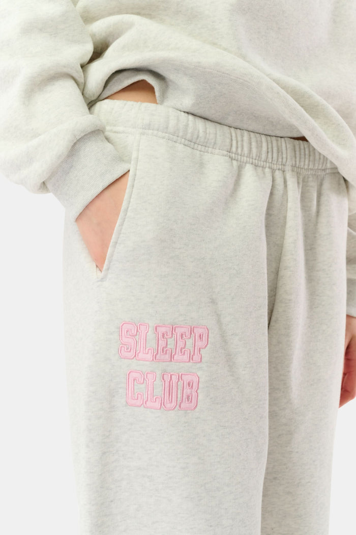A woman stands against a white background wearing light gray Roller Rabbit Sleep Club Bea Sweatpants and a matching sweatshirt with "SLEEP CLUB" in pink, styled with pink and black Adidas sneakers.