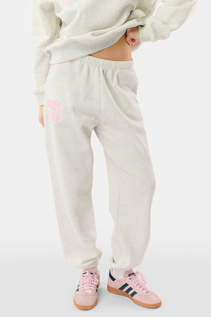 A woman stands against a white background wearing light gray Roller Rabbit Sleep Club Bea Sweatpants and a matching sweatshirt with "SLEEP CLUB" in pink, styled with pink and black Adidas sneakers.