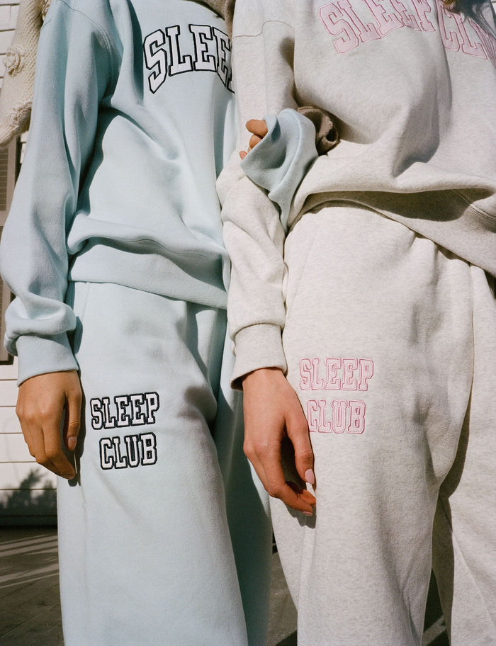 Model wears light blue Roller Rabbit Sleep Club Bea Sweatpants with “SLEEP CLUB” on the left leg, a matching sweatshirt, and white sneakers, against a plain background. Cozy looped terry fabric ensures comfort.