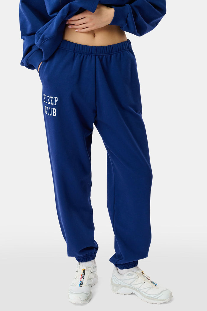 A young woman faces forward in a matching blue hoodie and Roller Rabbit Sleep Club Bea Sweatpants with "SLEEP CLUB" printed in white, paired with white sneakers. The hoodie has a small white graphic.